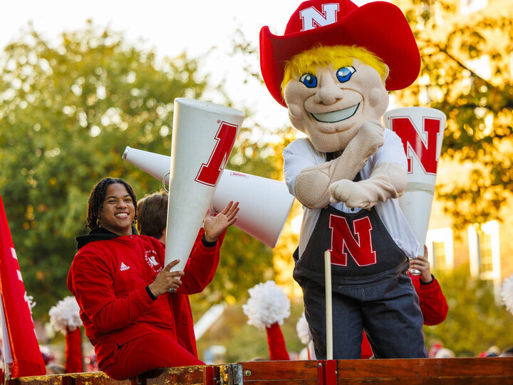 Herbie Husker and Spirit Squad member Amani Mfinanga cheer from a tractor-pulled wagon in the 2023 homecoming parade. [Craig Chandler | University Communication and Marketing]