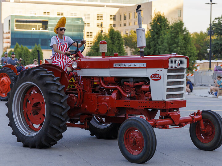 Student wears red and while striped overalls and a Nebraska Cornhead Hat while driving a vintage Farmall tractor. [Mike Jackson | Student Affairs] 