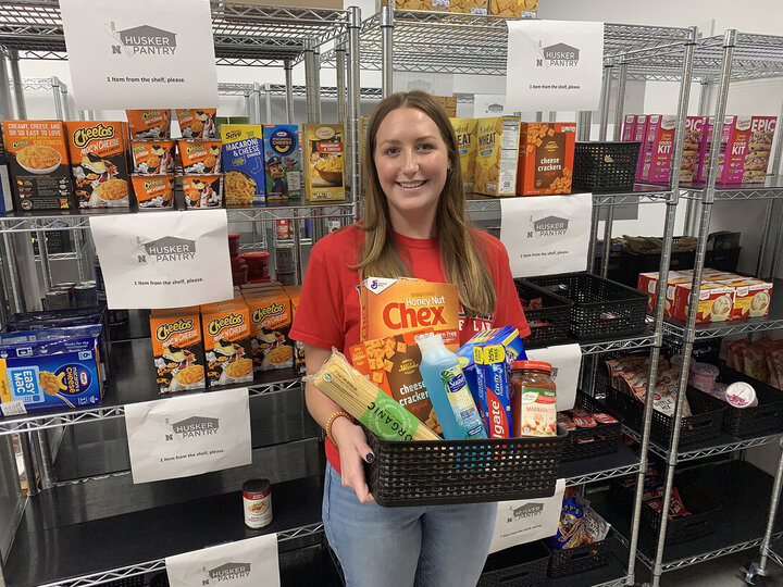 Woman holding a basket of groceries in front of pantry shelves stocked with food items.